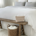Small beige luxury padded device bag on wooden bench beside bed with white and grey bedding