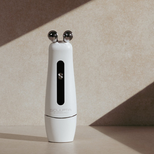 White Sculpta by Déesse PRO facial massage device with dual metal spheres on beige surface